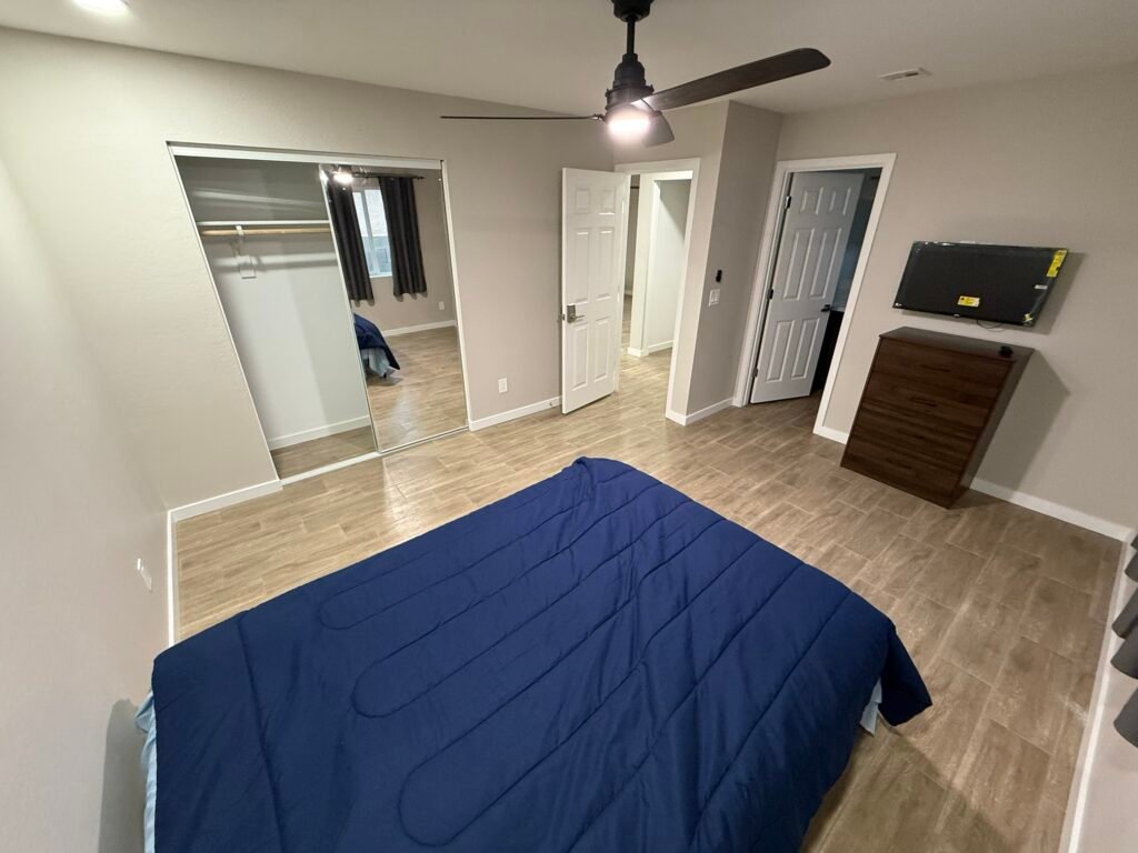 Large private room with full-size bed, dresser, wall-mounted TV, ceiling fan and mirrored closet at a Housing Done Wright home in Phoenix